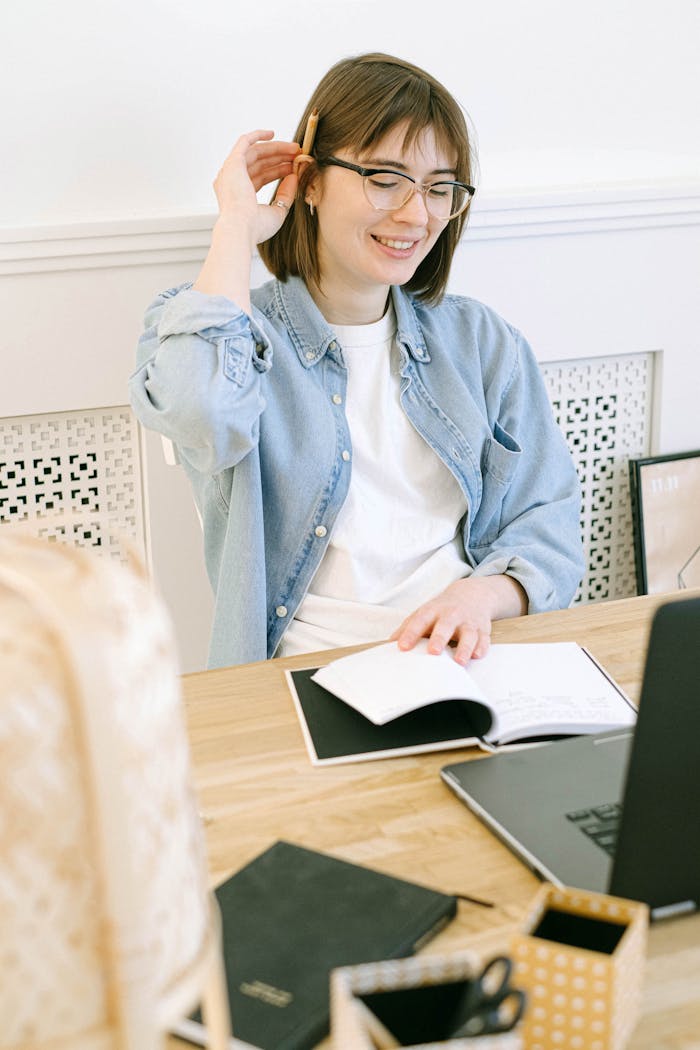 Smiling woman in casual attire working at home office with a laptop and notebook.