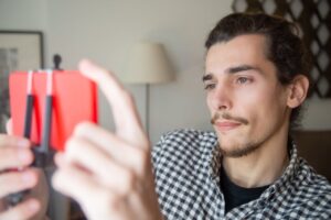 A young bearded man in a checkered shirt live streaming indoors using a smartphone on a tripod.